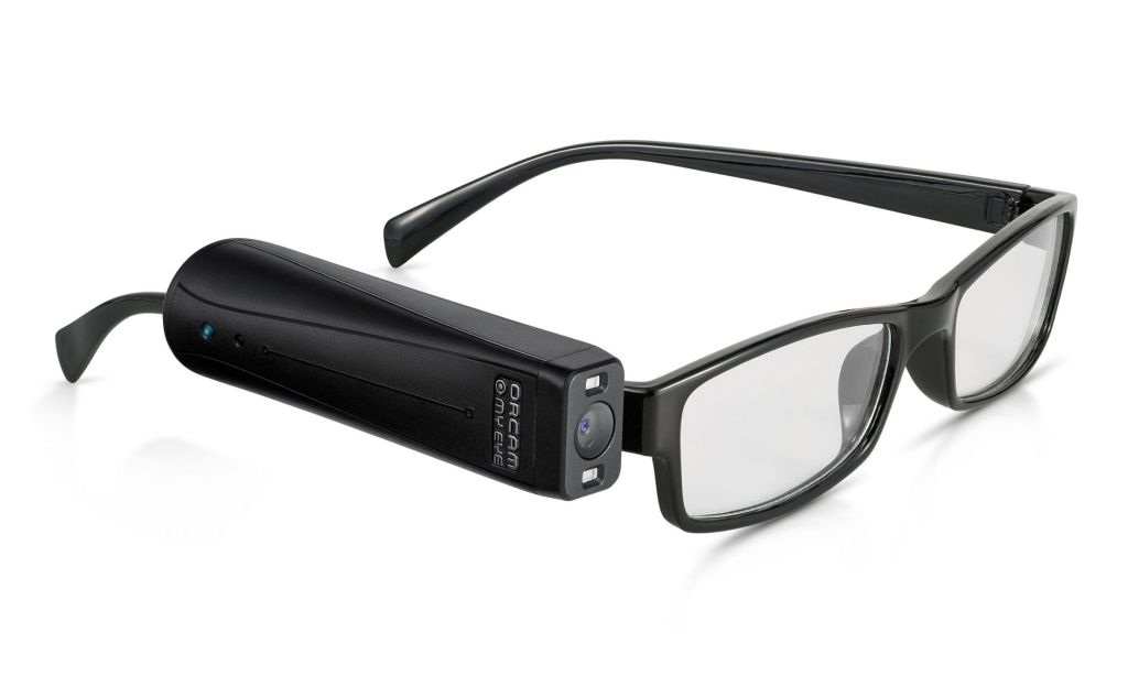 A photo of a pair of black-framed glasses with the Orcam MyEye clipped to the side. The Orcam MyEye is black and sleek, about the size and shape of a lipstick tube, with a sensor on the outside and a camera lens on the front.